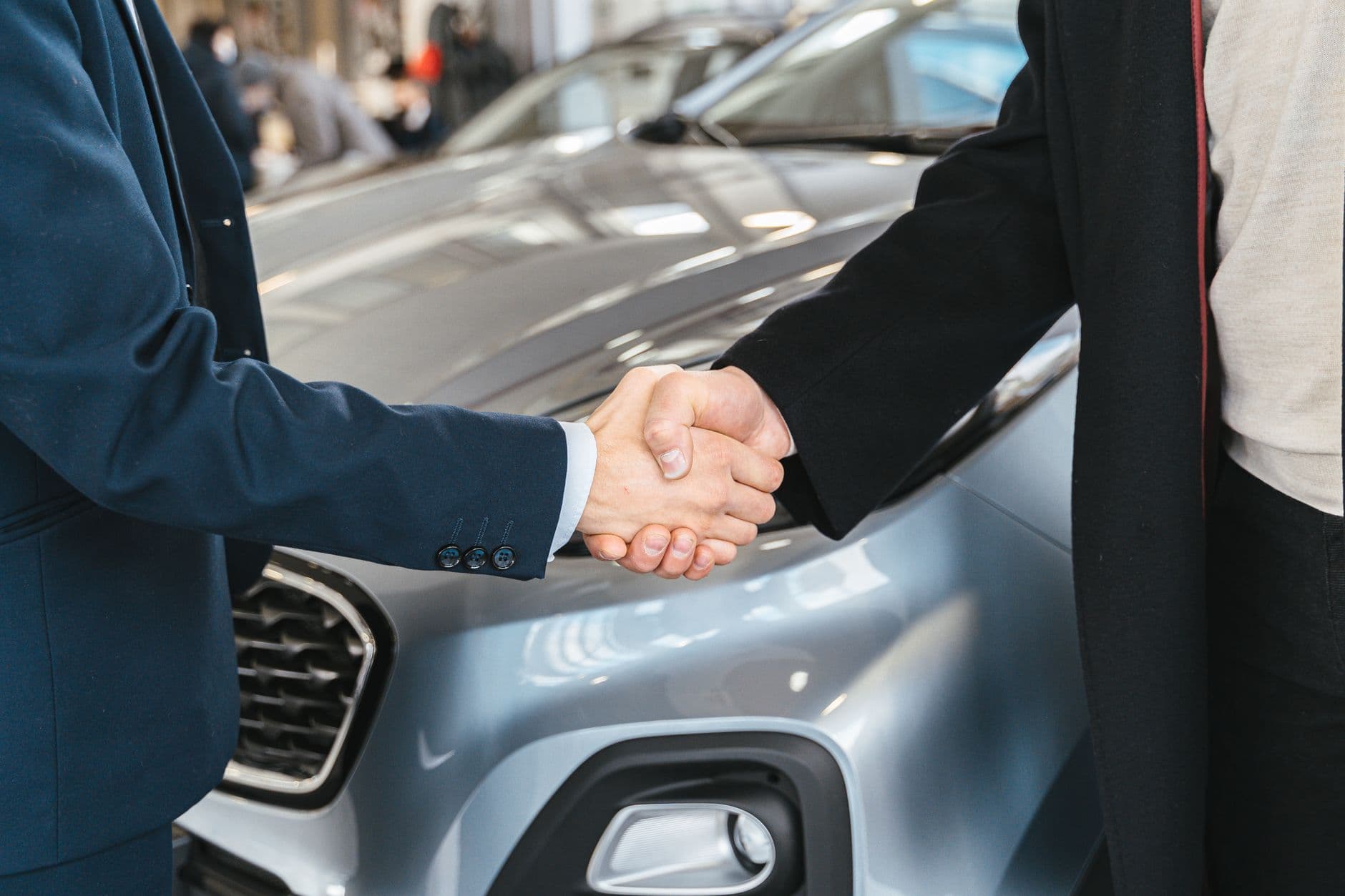 Dos personas dándose la mano frente al capó de un carro gris, cerrando el negocio de la venta