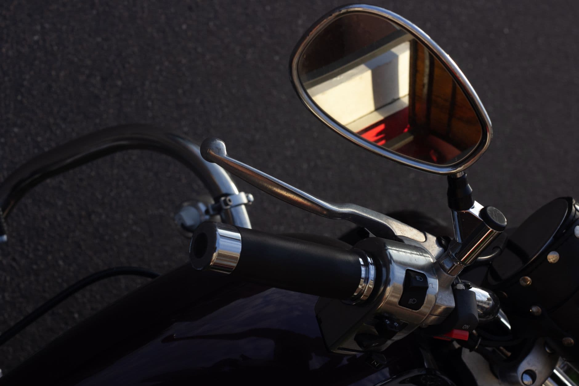 Primer plano del manubrio de una moto: el espejo retrovisor, la maneta del freno y el mando de las luces, con el asfalto de fondo