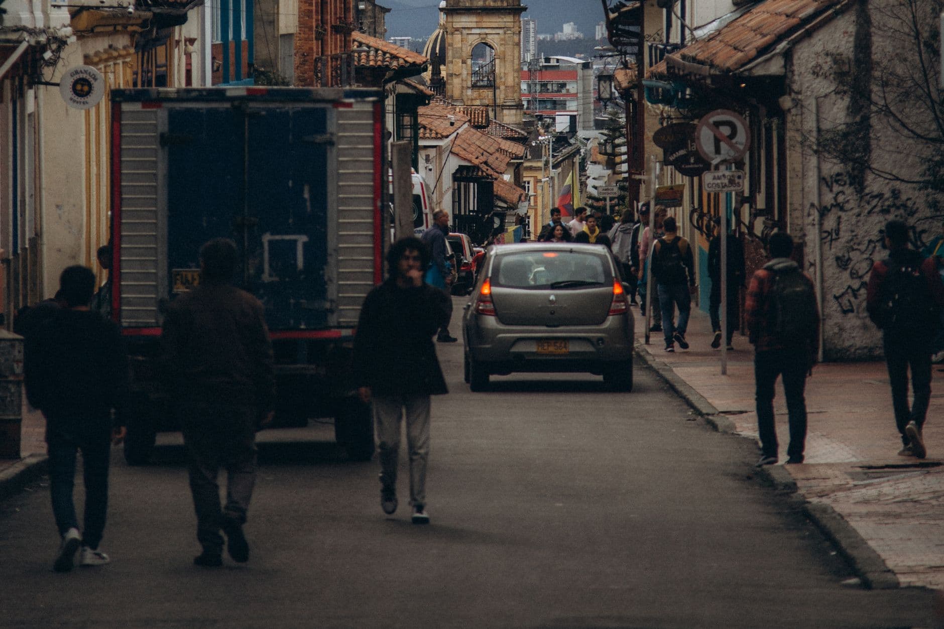 Carro particular gris circulando por una calle de La Candelaria en Bogotá, entre peatones y con una bandera de Colombia al fondo